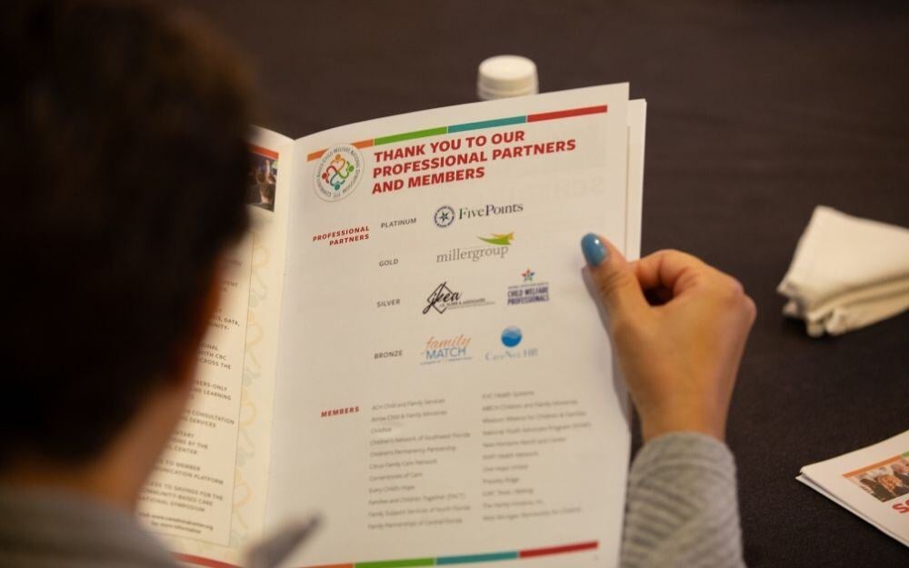 A closeup view of the National Center for Community-Based Child Welfare’s 2024 Symposium booklet showing all the logos of its partners, members, and sponsors with an attendee reading it in the foreground.